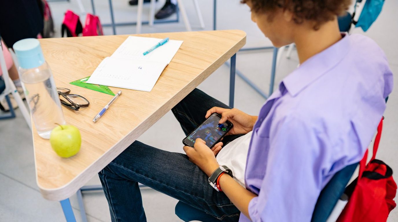 Estudante sentado à mesa estudando com material de apoio e celular.