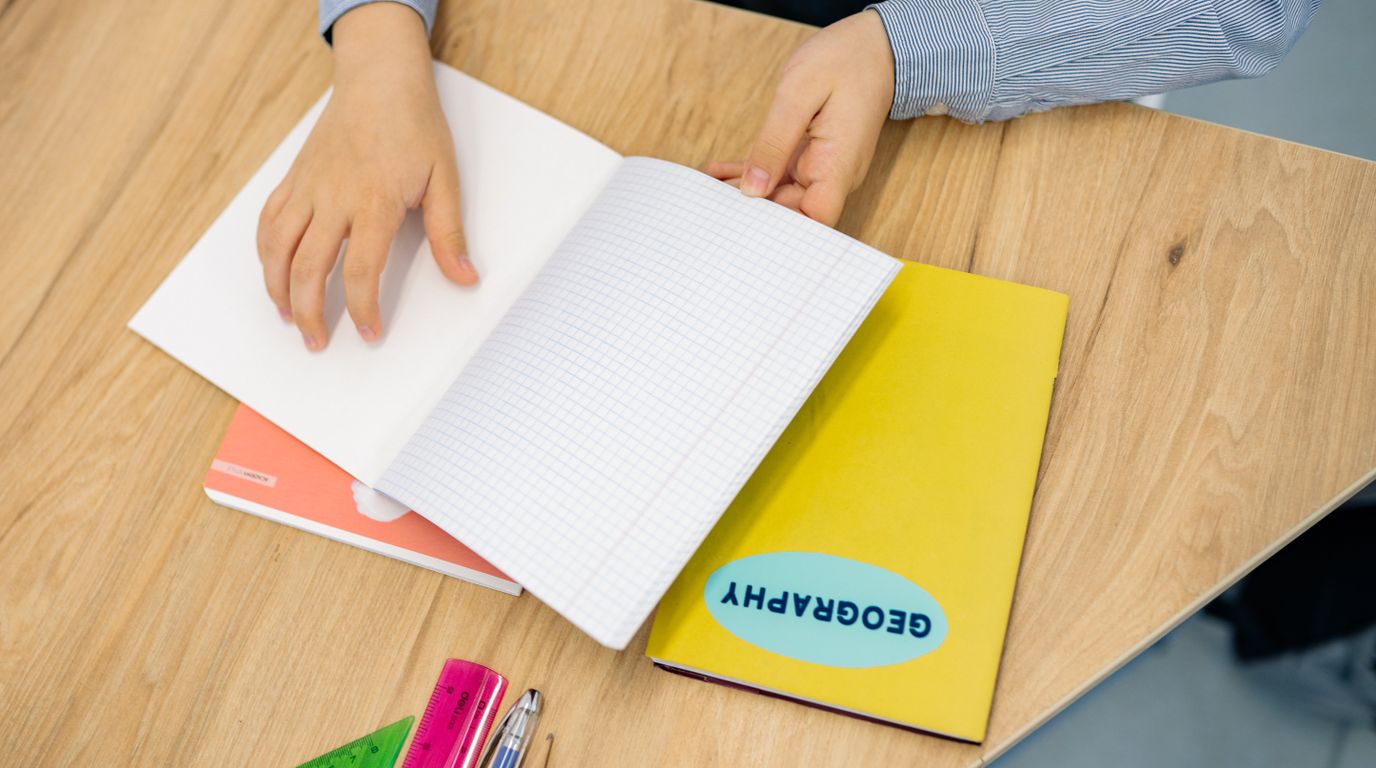 Mãos folheando um caderno quadriculado aberto sobre a mesa.